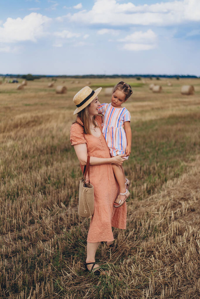 A young mother in a long dress and a straw hat leads a little daughter by the hand. Both walk across a field of harvested wheat. Against the background of a bale of straw - Photo, Image