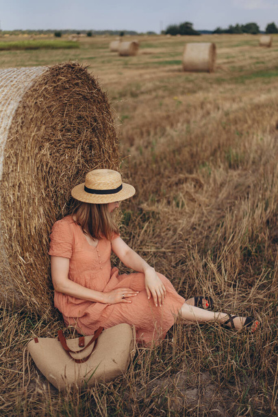 長いドレスの美しい女の子と長い髪の笑顔は収穫された小麦の畑のわらに覆われたかわいい座っています. ストローハットガール - 写真・画像