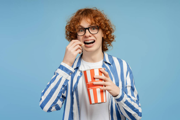 Rozradowany rudy chłopiec trzymający wiadro z popcornem, jedzący przekąski oglądający film na niebieskim tle. Kino, koncepcja reklamy  - Zdjęcie, obraz
