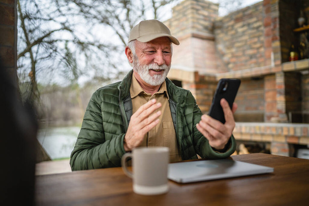 jeden mężczyzna starszy kaukaski mężczyzna z szary broda i czapka siedzieć na zewnątrz na wiosnę lub jesień dzień szczęśliwy uśmiech używać telefon komórkowy dla wideo połączenie skopiować przestrzeń prawdziwi ludzie - Zdjęcie, obraz
