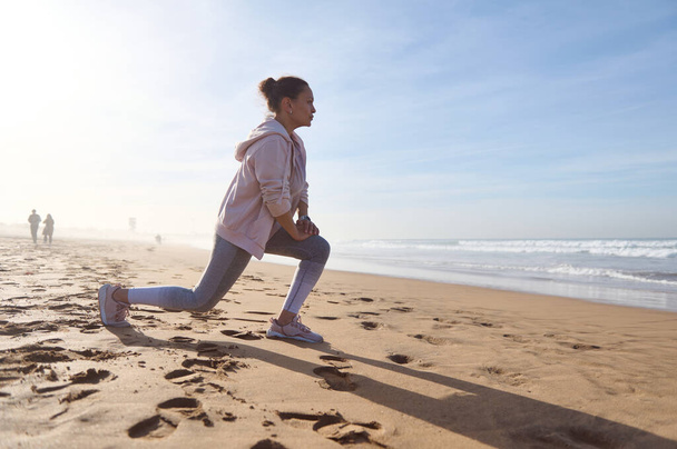 Mulher esportiva ativa em activewear exercício na praia de areia, fazendo lunges enquanto aquece seu corpo, alongando os músculos das pernas durante o treino matinal. Gente. Desporto. Conceito de estilo de vida saudável ativo - Foto, Imagem