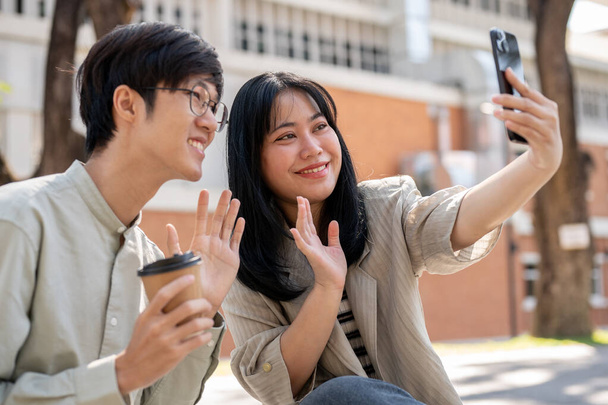 Zwei gut gelaunte asiatische Freunde fotografieren in der Mittagspause gemeinsam mit einem Smartphone beim Chillen im Stadtpark. - Foto, Bild