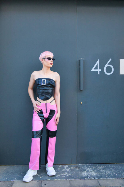Giovane ragazzo gay con i capelli rosa e make-up e vestito di rosa e nero è appoggiato contro una porta del garage nero. Il ragazzo indossa occhiali da sole e sta facendo pose diverse. Concetto di diritti LGBTQ - Foto, immagini