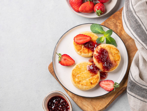 Cottage kaas, cheesecakes of syrniki bestrooid met bessensaus op een snijplank op een lichte achtergrond met jam, aardbei en servet. Het concept van een voedzaam en gezond ontbijt. Bovenaanzicht - Foto, afbeelding