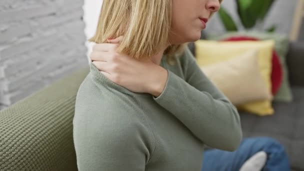Una mujer rubia con el pelo corto experimenta dolor de cuello en casa en un sofá, retratando el malestar en el interior. - Imágenes, Vídeo