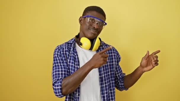 Γελώντας αφροαμερικάνος άνδρας με αυτοπεποίθηση έκφρασης εμπιστοσύνης, φορώντας γυαλιά ασφαλείας, στέκεται και δείχνει προς την πλευρά με τα δύο χέρια στο φωτεινό κίτρινο απομονωμένο φόντο - Πλάνα, βίντεο