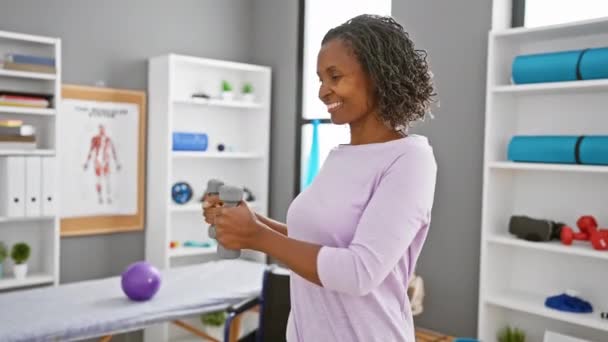 Uma mulher afro-americana sorridente se exercita com halteres em uma clínica de fisioterapia bem equipada, refletindo cuidados de saúde e estilo de vida ativo. - Filmagem, Vídeo