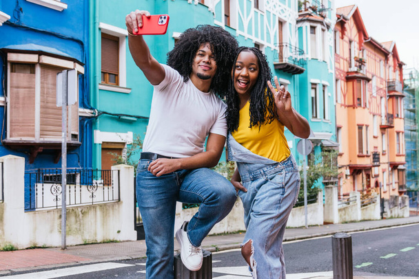 Latino friends posing for a lively selfie on a city street, surrounded by colorful houses, radiating happiness and style. - Photo, Image