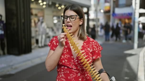 Красивая латиноамериканка радостно ест хрустящие, вкусные чипсы на палочке на улице Такешита, токё - путешествующая любительница нездоровой пищи, спортивные очки и веселую улыбку в японском городке - Кадры, видео