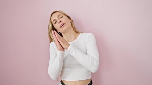 Dreamy blonde woman, young and tired, posing asleep standing with hands together, a subtle smile on her face, her eyes closed, oozing positive energy over an isolated pink background. - Footage, Video