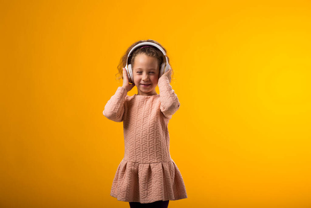 イエローバックグラウンドでヘッドフォンで音楽を楽しむピンクのドレスを着た笑顔の少女の肖像. ライフスタイルとリースコンセプト - 写真・画像