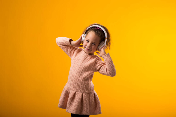 Portrét usměvavé dítě dívka na sobě růžové šaty těší hudbu ve sluchátkách na žlutém pozadí. Koncept životního stylu a volného času - Fotografie, Obrázek