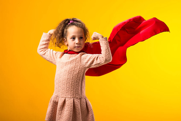 Portrait de petite fille super-héros dans un manteau rouge montrant geste tendu sur fond jaune. Concept de victoire et de succès. - Photo, image
