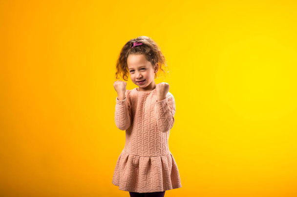 Portrét usměvavé dívky, která ukazuje úspěch na žlutém pozadí. Koncept štěstí - Fotografie, Obrázek