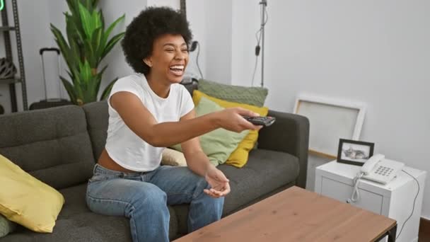 Mujer sonriente con el pelo rizado sosteniendo un control remoto en una acogedora sala de estar - Metraje, vídeo