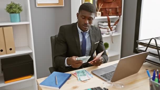 Egy afrikai üzletember egy irodában számolja a kínai jüant, miközben különböző érzelmeket fejez ki az asztalánál egy laptoppal, számológéppel és jegyzetfüzetével.. - Felvétel, videó