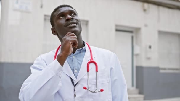 Przemyślany Afrykanin w płaszczu lekarza ze stetoskopem stoi przed kliniką.. - Materiał filmowy, wideo
