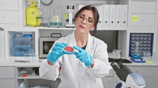 Μια στοχευμένη γυναίκα επιστήμονας στο εργαστήριο παλτό εξετάζει ένα δείγμα σε ένα σύγχρονο εργαστηριακό περιβάλλον, απεικονίζοντας τον επαγγελματισμό και την προσοχή στη λεπτομέρεια. - Πλάνα, βίντεο
