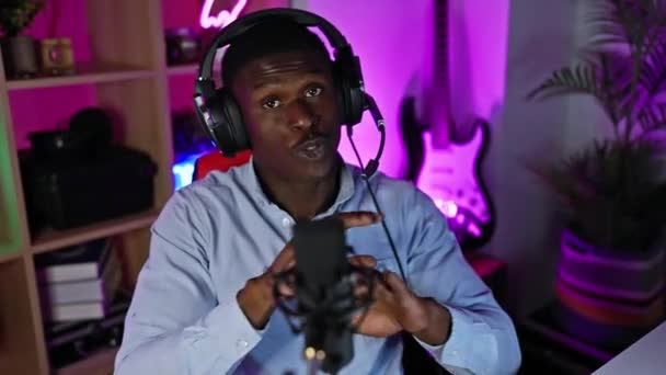 Egy afro-amerikai férfi podcasting fejhallgatóval és mikrofonnal egy színes játékteremben éjszaka. - Felvétel, videó