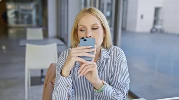 Een blonde vrouw in gestreept shirt maakt gebruik van smartphone in een outdoor cafe. - Video