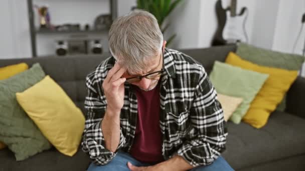 Ein nachdenklicher älterer Mann mit Brille und grauen Haaren sitzt zu Hause auf einem Sofa und schaut besorgt.. - Filmmaterial, Video
