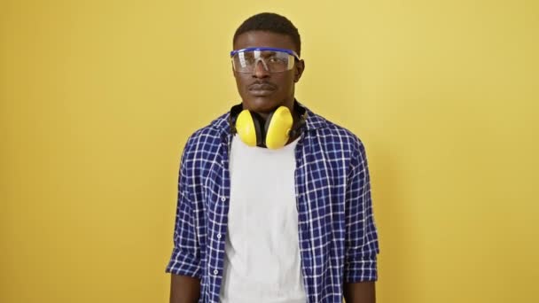 Selbstbewusster afrikanisch-amerikanischer Mann mit ernstem Gesichtsausdruck, der eine Schutzbrille aufsetzt. Er steht perfekt ausbalanciert und verkörpert einen natürlichen, einfachen Blick. eine isolierte Gestalt vor gelbem Hintergrund. - Filmmaterial, Video