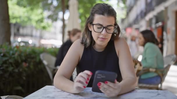 Χαμογελώντας όμορφη ισπανίδα γυναίκα, φορώντας γυαλιά, κάθεται άνετα σε ένα τραπέζι καφέ, ευτυχώς χρησιμοποιώντας το smartphone της σε εξωτερικούς χώρους - Πλάνα, βίντεο