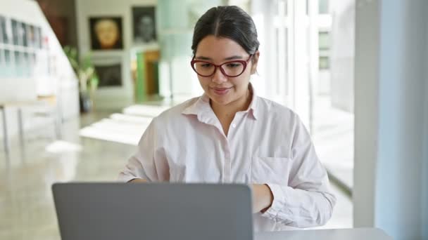 Νεαρή ισπανόφωνη γυναίκα με γυαλιά που εργάζεται σε φορητό υπολογιστή στο σύγχρονο γραφείο με θολή εσωτερικό υπόβαθρο. - Πλάνα, βίντεο