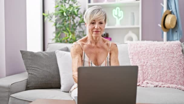 Kypsä nainen, jolla on lyhyet harmaat hiukset, rentoutuu olohuoneen sohvalla sulkemalla kannettavan tietokoneen, joka kuvaa tunteen saavutuksesta ja vapaa-ajasta kotona.. - Materiaali, video