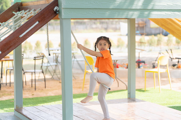 Jong meisje dat op een touwladder speelt. Een klein meisje in een oranje shirt klimt op een touwladder op een speelplaats, terugkijkend met concentratie. Baby meisje kinderen activiteit concept. - Foto, afbeelding