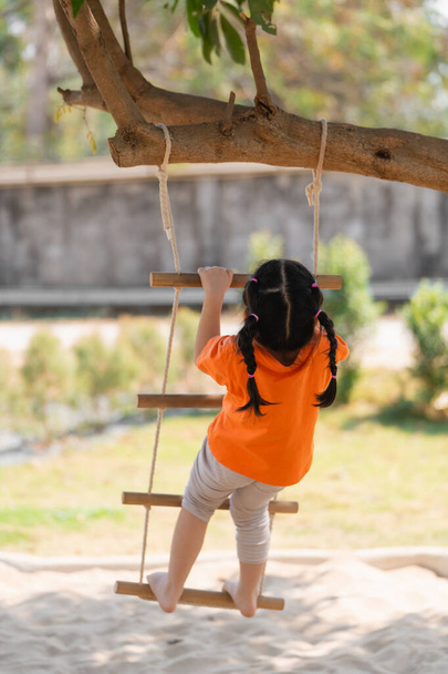 Visszanézni. Egy fiatal lány kötéllétrán játszik. Egy narancssárga inges kislány felmászik egy kötélhágcsóra a játszótéren, koncentráltan. Baby girl kids tevékenység koncepció. - Fotó, kép