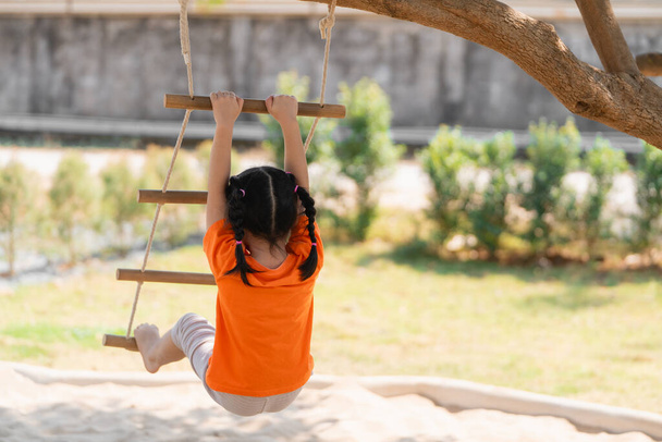 Vue de derrière. Jeune fille jouant sur une échelle de corde. Une petite fille en chemise orange grimpe une échelle de corde sur une aire de jeux, avec concentration. Bébé fille enfants concept d'activité. - Photo, image