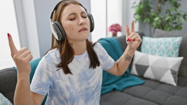 Eine junge Frau genießt Musik mit Kopfhörern in einem gemütlichen Wohnzimmer und gestikuliert mit ihren Händen. - Filmmaterial, Video