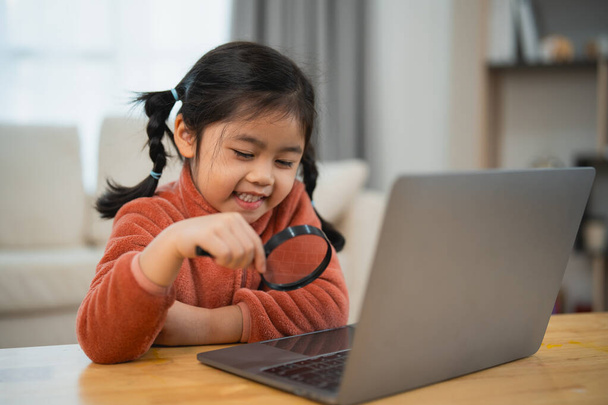 Nieuwsgierig meisje verkennen met vergrootglas en laptop. Glimlachend jong meisje met vlechtjes met een vergrootglas om naar een laptopscherm te kijken, nieuwsgierig en blij. Onderwijs voor kinderen concept. - Foto, afbeelding