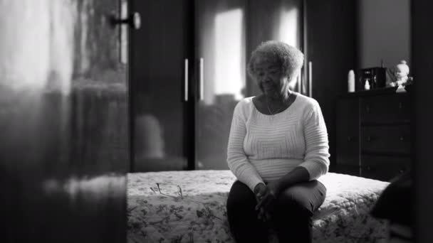 Eine nachdenkliche schwarze ältere Dame sitzt in dramatischem Schwarz-Weiß am Krankenbett. Nachdenkliche Seniorin in bescheidener Einsamkeit zu Hause, nachdenklicher Blick, der über das Leben nachdenkt - Filmmaterial, Video
