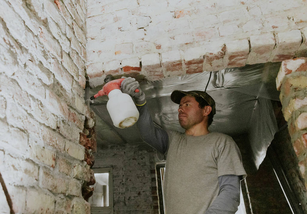 Un jeune bel homme brune caucasien en vêtements décontractés se tient sur un escabeau et pulvérise de l'eau à partir d'un vaporisateur sur des briques dans une porte sous le plafond dans une vieille maison, vue rapprochée du bas. - Photo, image