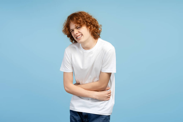 Portret uwieczniający kręconego rudego nastolatka z aparatem na zębach, gryzącego i trzymającego brzuch, ustawionego na niebieskim tle. Reprezentuje ból brzucha. Koncentracja na koncepcji opieki zdrowotnej - Zdjęcie, obraz