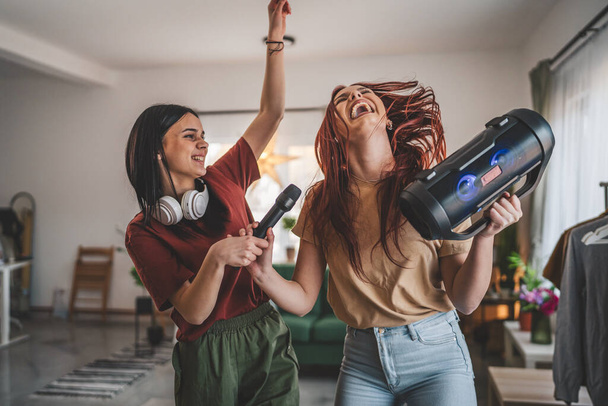 Two women young caucasian friends or sisters have fun at home females dance and sing karaoke hold microphone listen to the music happy smile joyful rhythm real people copy space - Photo, Image