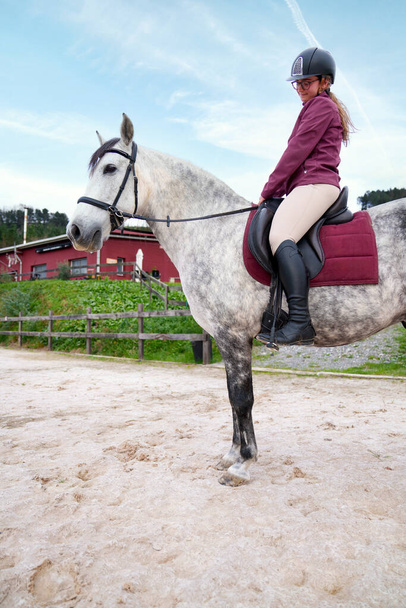 Fiducioso giovane equestre sorridente mentre montato su un cavallo bianco inzuppato, con una stalla rossa sullo sfondo. - Foto, immagini
