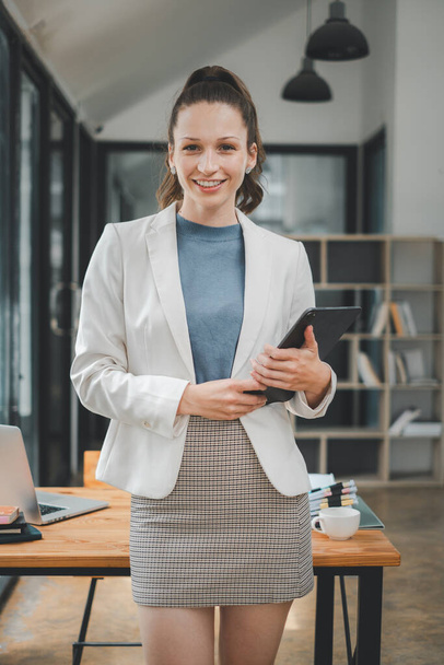 Business woman koncepció, professzionális fiatal nő egy elegáns fehér blézer mosolygós, kezében egy tabletta egy kortárs irodai környezetben. - Fotó, kép