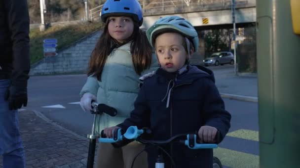 Kleine jongen en meisje wachten om de straat over te steken op voetgangersoversteekplaats met beschermende helmen op de top van scooter en fiets dragen jassen tijdens de koude dag. Verantwoordelijke kinderen wachten op groen licht - Video