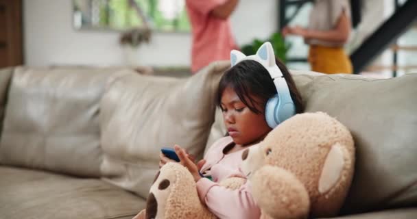 Дитина, телефон і навушники з батьками сперечаються, не погоджуються або розлучаються на дивані вітальні вдома. Маленька дівчинка або дочка грають на мобільному смартфоні з плюшевим ведмедем у сімейному конфлікті. - Кадри, відео