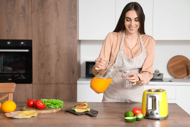 Νεαρή όμορφη ευτυχισμένη γυναίκα με νόστιμο σάντουιτς ρίχνει χυμό πορτοκαλιού σε ποτήρι στην κουζίνα - Φωτογραφία, εικόνα