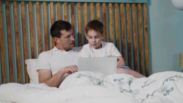 A friendly father and son in a joyful mood lie on the bed in the bedroom with a laptop, smile, laugh and talk. A positive dad with child watch a funny movie or cartoon and discuss it. The concept of - Footage, Video