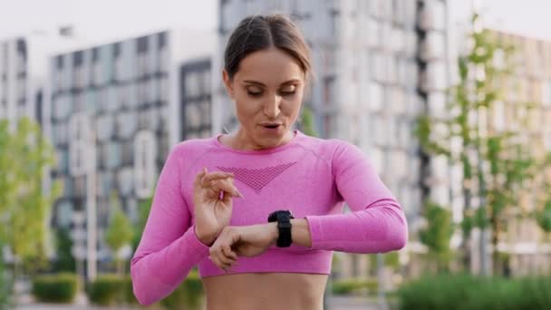A young charming girl jogger outdoors in the city park among modern residential highrise buildings. A confident sportswoman performs a sprinting, set a record of running, checks the time on the watch - Footage, Video