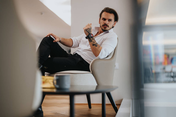 Αυτοπεποίθηση νεαρός άνδρας με τατουάζ κάθεται άνετα σε μια καρέκλα γραφείου, αποπνέοντας δροσερό επαγγελματισμό και μοντέρνο στυλ. - Φωτογραφία, εικόνα