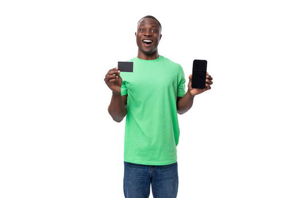 Açık yeşil tişörtlü ve kot pantolonlu genç, bakımlı Afrikalı bir adam bir kredi kartı ve bir akıllı telefon gösteriyor.. - Fotoğraf, Görsel