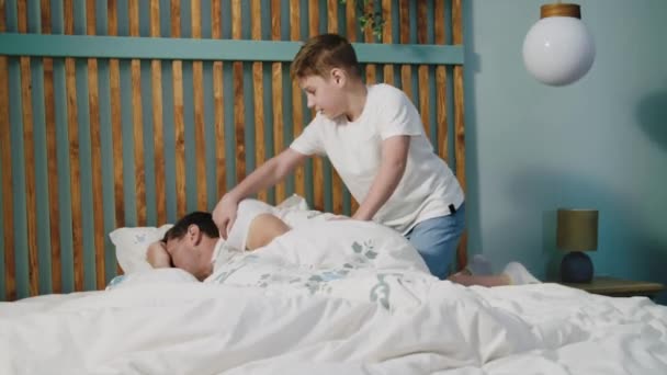 A son enters into the bedroom to sleeping father, wakes him up and shakes by the shoulder. A positive family of dad and child lies on the bed, laughs, plays and has fun. The concept of happy childhood - Footage, Video