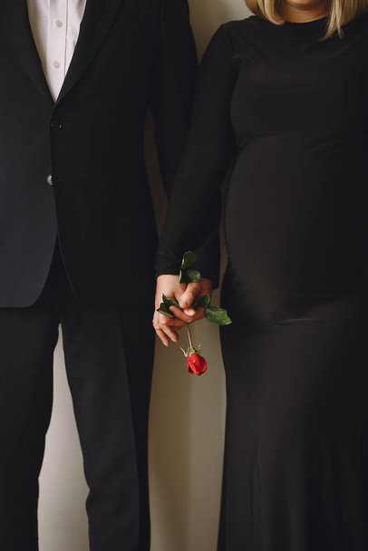 妊娠中の女性は黒いドレスを着て,夫は赤いバラの花を妊娠中の腹の近くで手に持っています. 愛のコンセプト。 ファミリーコンセプト - 写真・画像