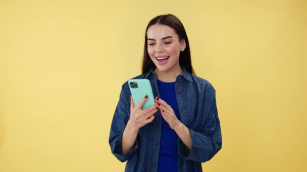 Stunning female brunette with shocked facial expression looking on mobile phone and pointing on screen. Casually dressed woman smiling at camera while recommending great new application in studio. - Footage, Video